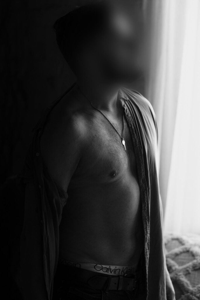 Male companion standing shirtless by the window, looking out thoughtfully