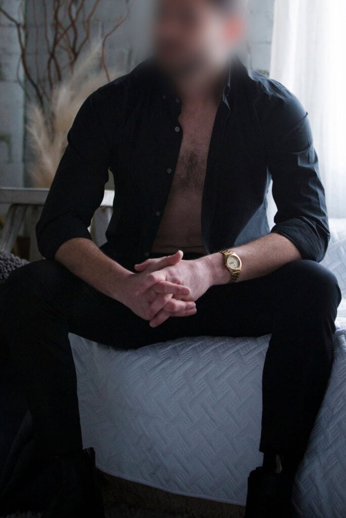 Companion seated casually, shirt open, relaxed and alluring.