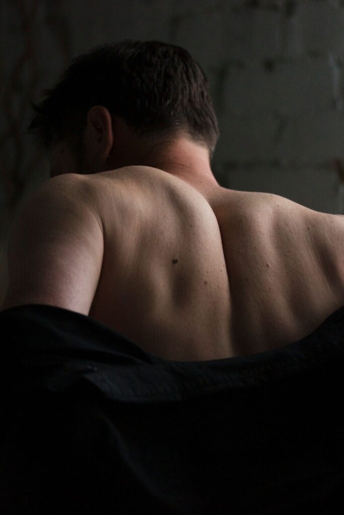 Male escort companion undressing, lifting shirt over head in dimly lit room